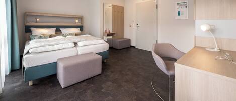 A modern hotel room with a large bed, a desk, and a chair. There is a door and a small table with a lamp.