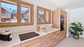A modern kitchen with wooden cabinetry, a built-in oven, and a large window offering a view of neighboring houses. - Le Framiré-appartement-4pers-Proche piste (Les Gets)