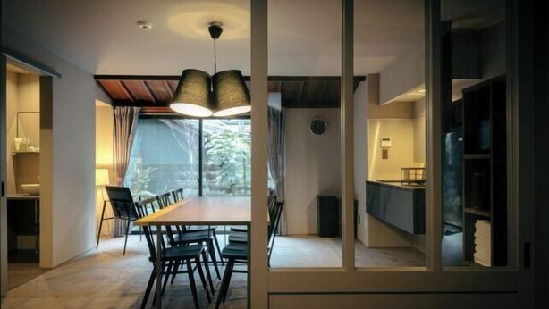 A dining area with a wooden table and chairs, a modern pendant light, and a view through a glass partition to a kitchen area. - SAKON UKON (Kanazawa)