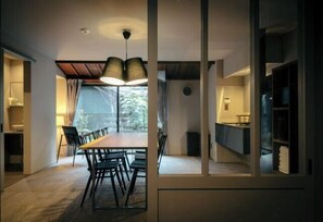 Un'area pranzo con un tavolo e sedie in legno, una lampada a sospensione moderna e vista, attraverso un divisorio in vetro, su una zona cucina.