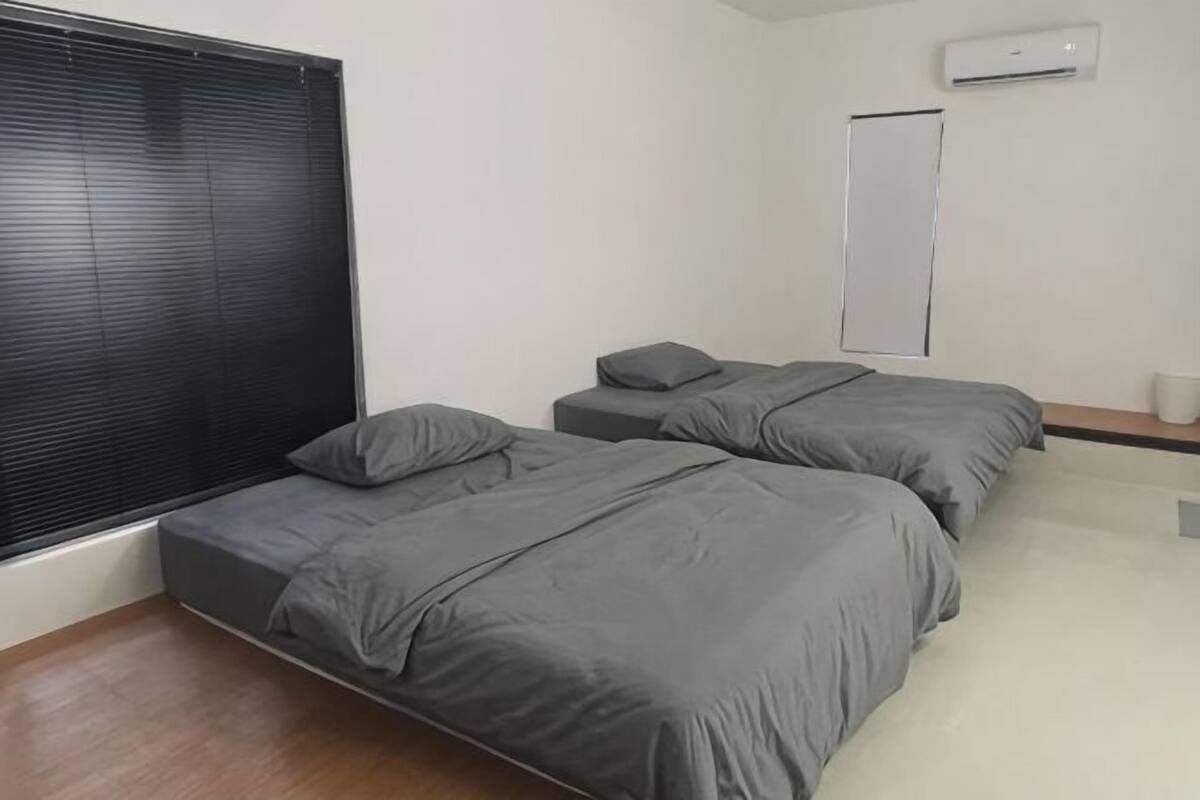 A bedroom with two beds, a window with blinds, an air conditioning unit, and a small bedside table.