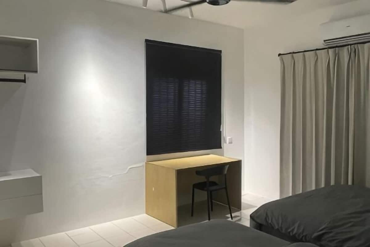 A bedroom with a bed, a desk, a chair, a window with blinds, and a curtain.