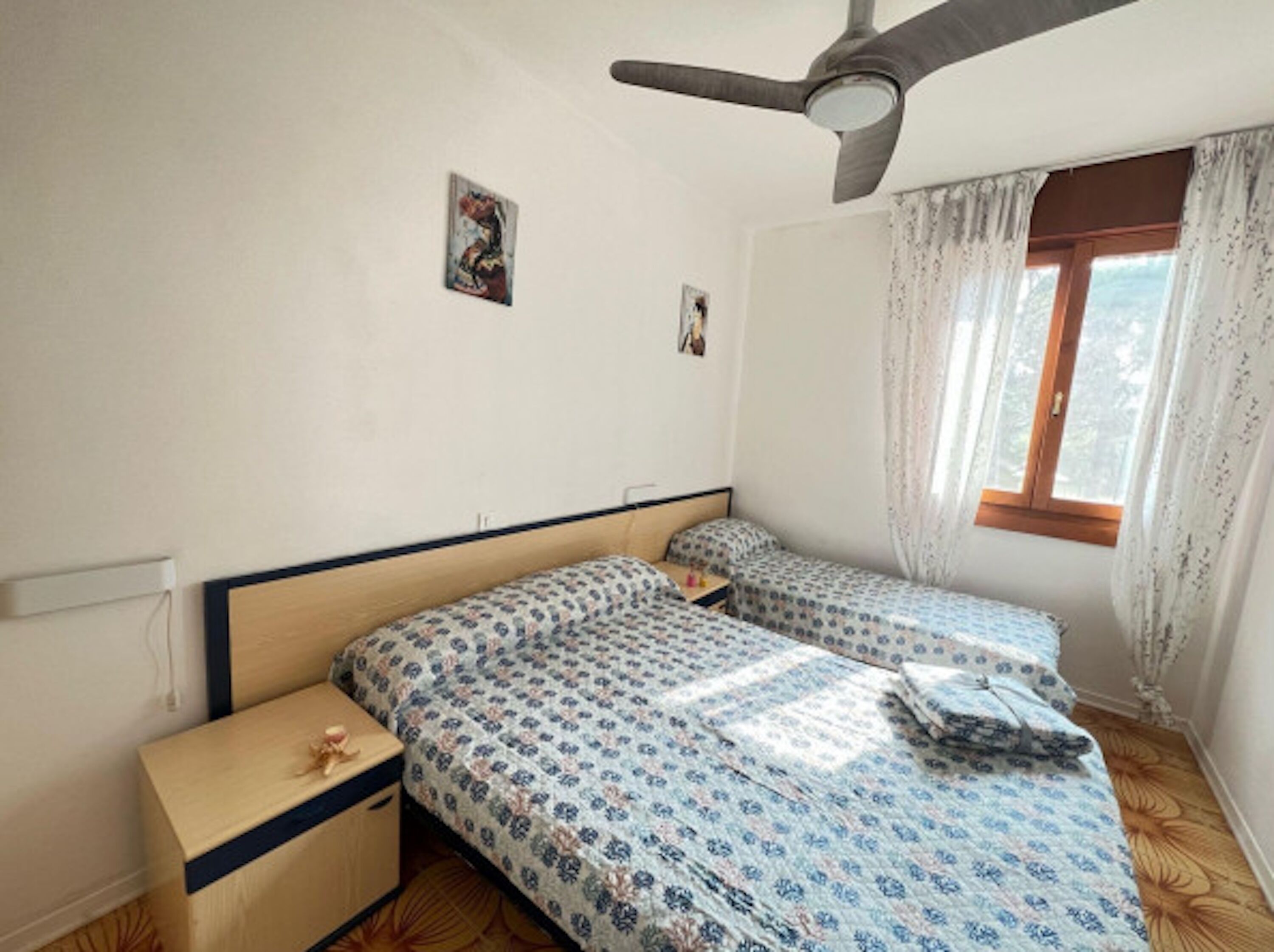 Un dormitorio con dos camas, un ventilador de techo, una ventana con cortinas y una mesita de noche pequeña.