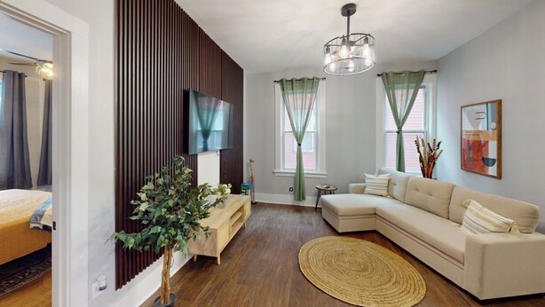 A modern living room with a sectional sofa, a flat-screen TV mounted on a wooden panel wall, a small side table, and a woven rug. - Spacious 6-bedroom townhome in charming Irvington with AC and Parking (Irvington)