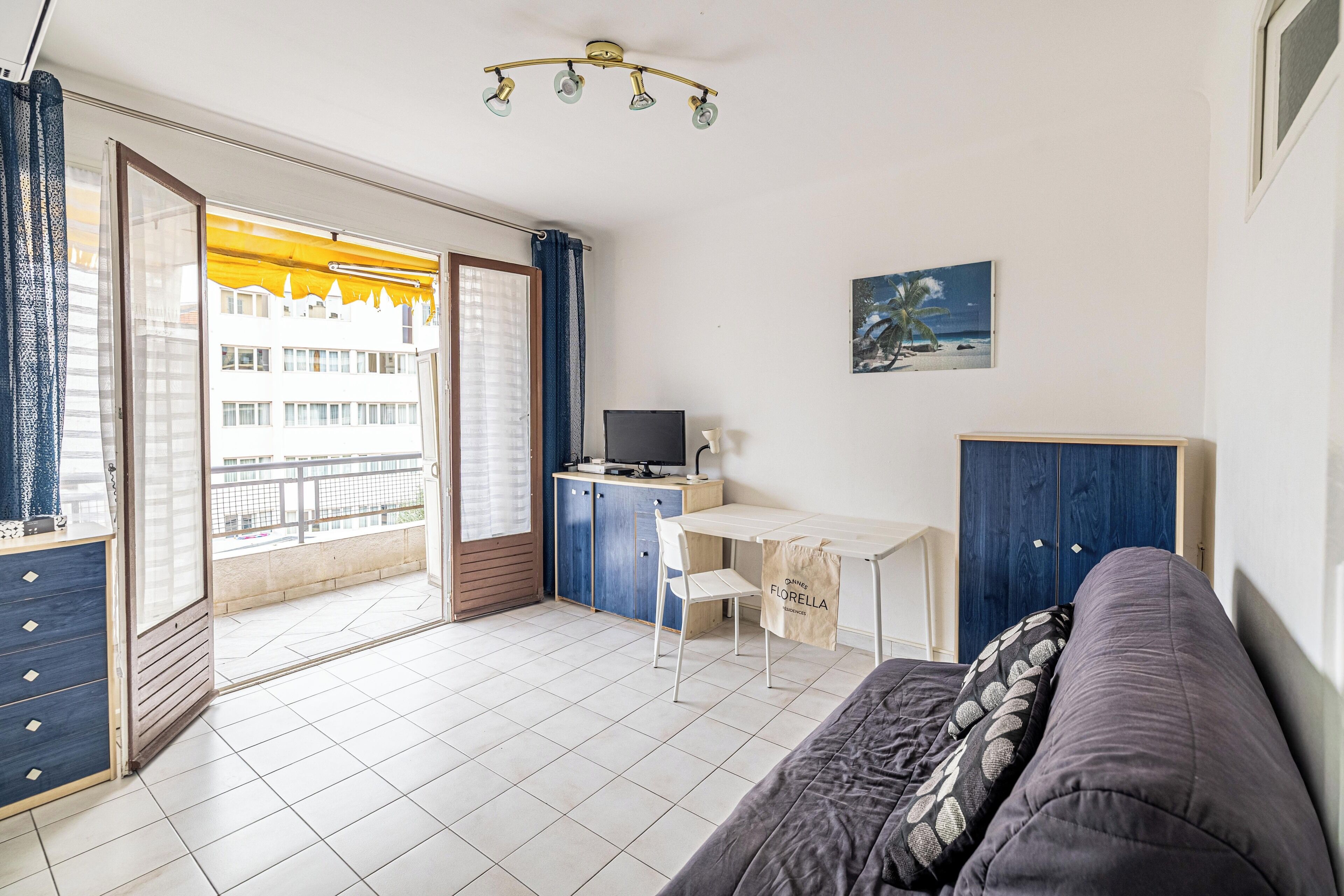 A room with a bed, a desk, a chair, a TV, and a balcony with a view of buildings.