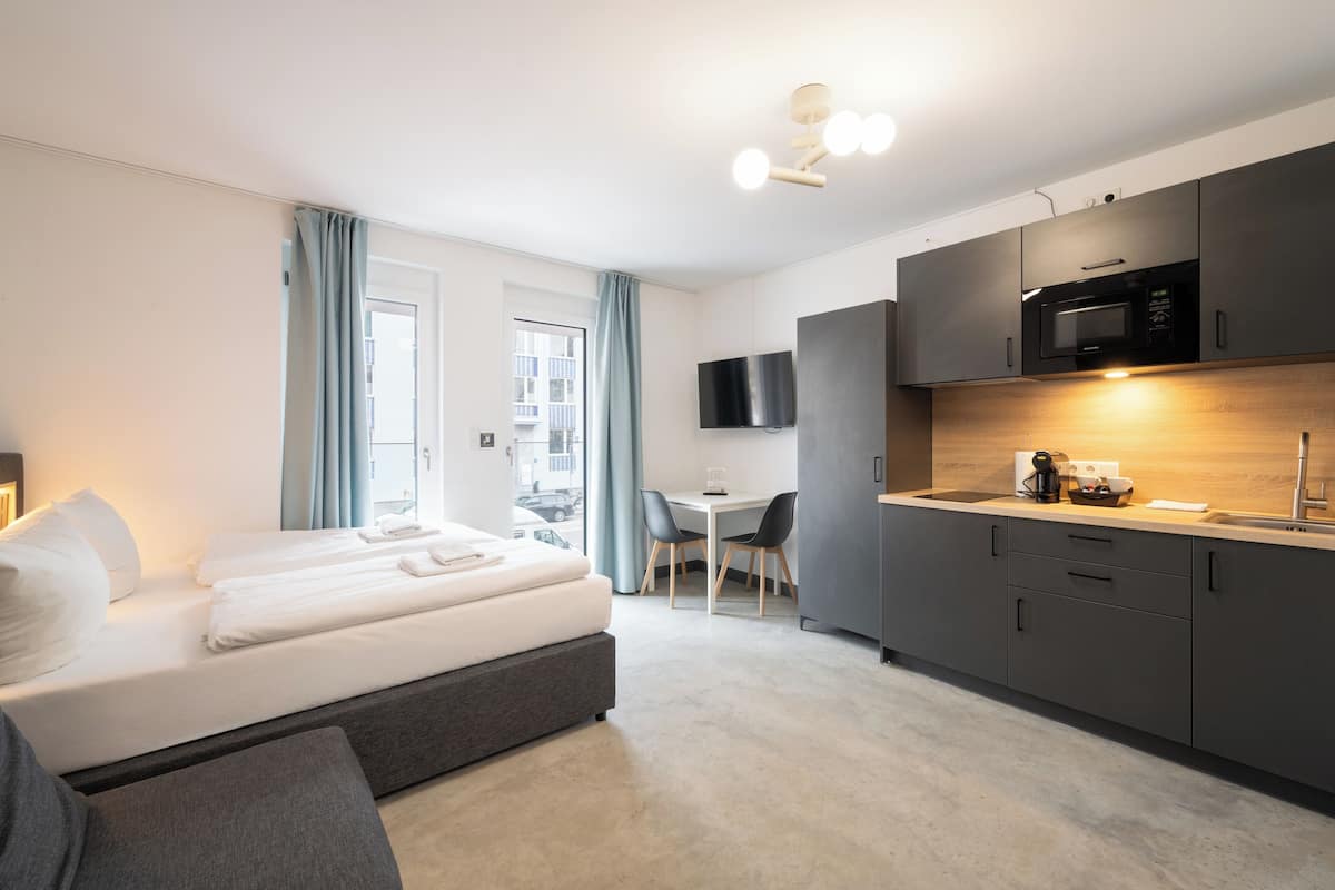 A modern hotel room with a bed, a desk, a kitchenette, and a TV.