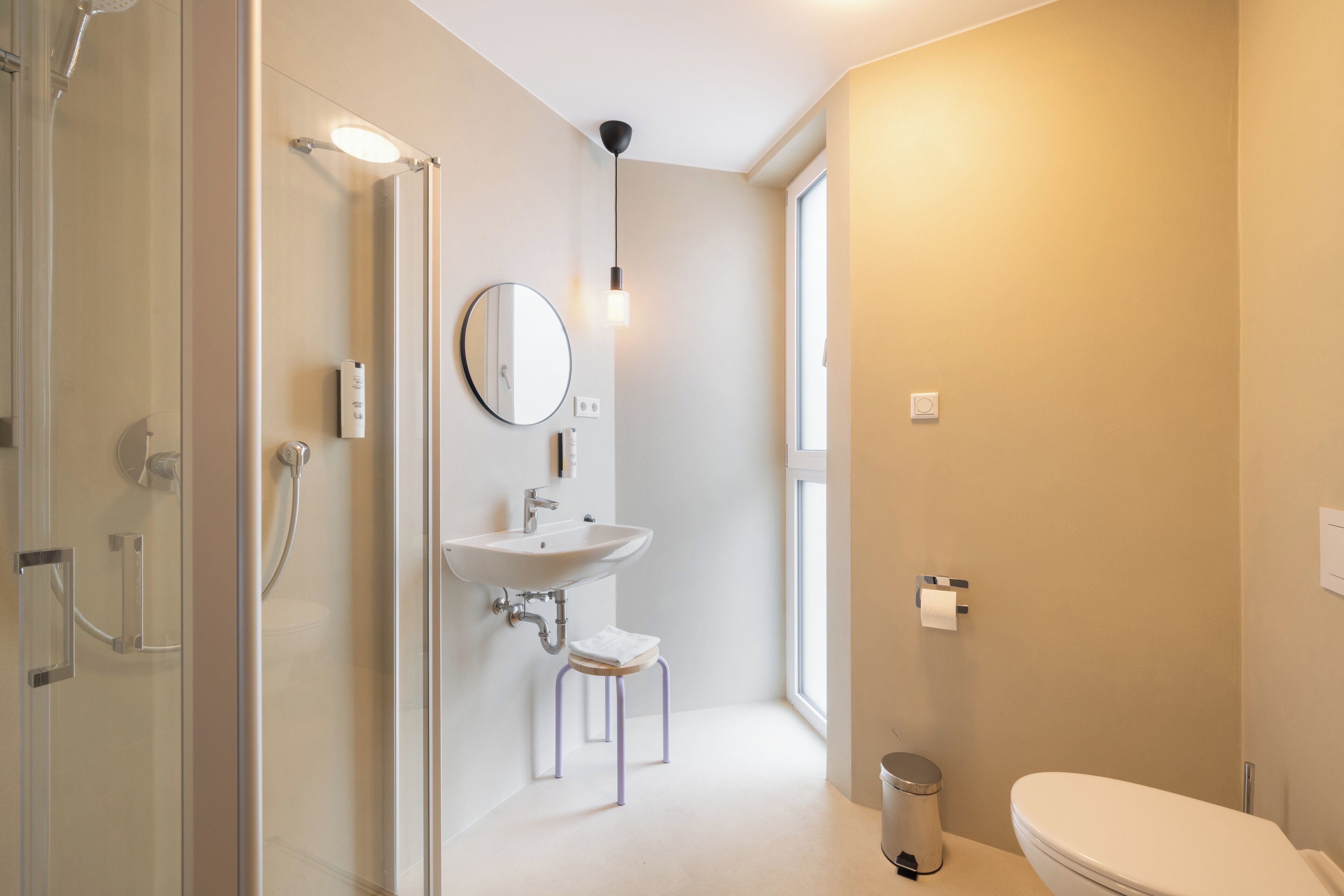 A modern bathroom with a glass shower enclosure, a pedestal sink, a toilet, and a window providing natural light.