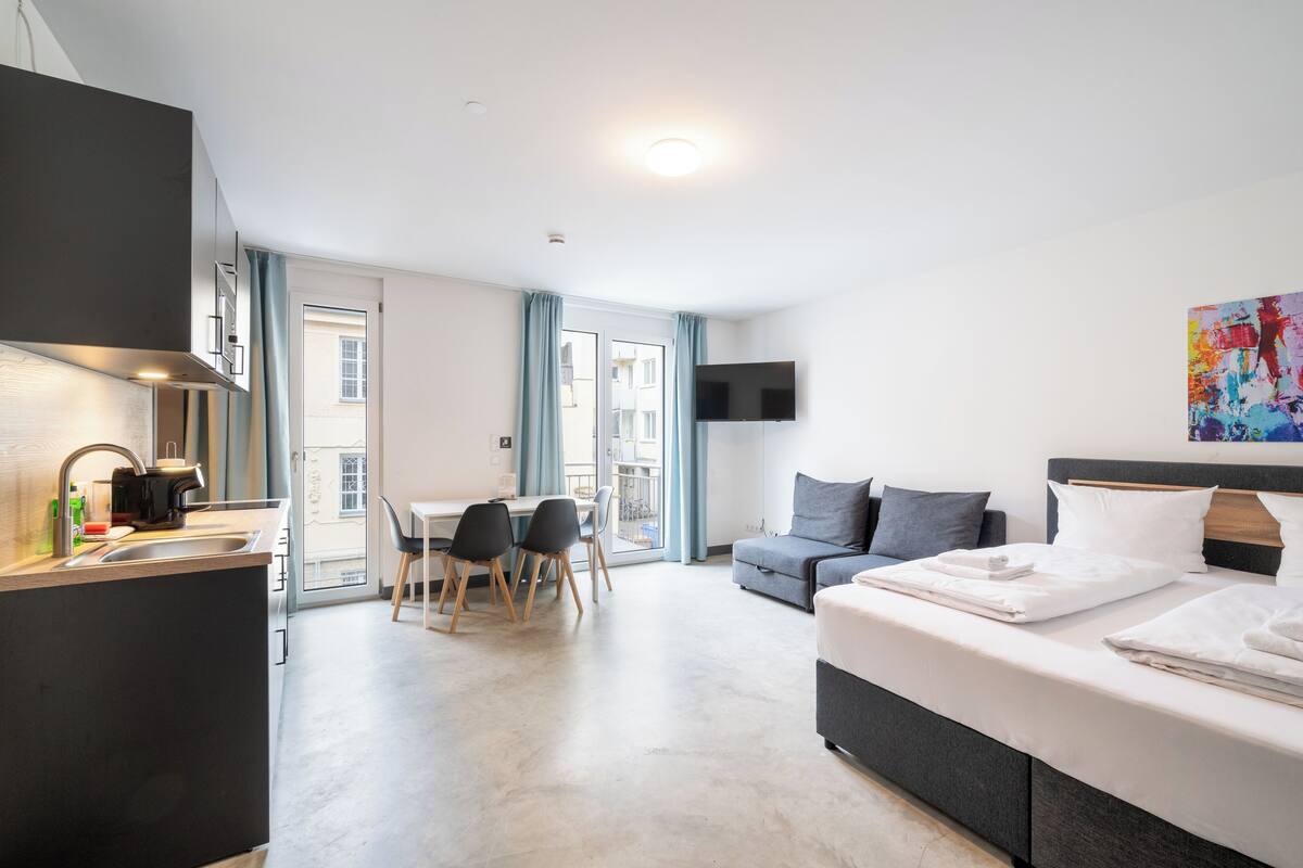 A modern hotel room with a kitchenette, a dining area, a sofa, and a bed.
