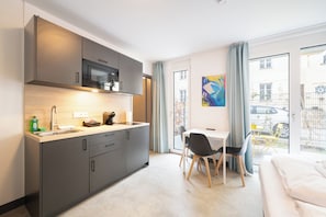 A modern kitchen with a microwave, sink, and cabinets, a dining area with a table and chairs, and a large window with a view of buildings and a car. - Homaris Apartments Therese (Munich)