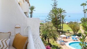 A balcony with a white wall, a cushioned bench, and a view of a swimming pool and beach. - Brand-new luxury with sea view  - Playa Real beachfront (Marbella)