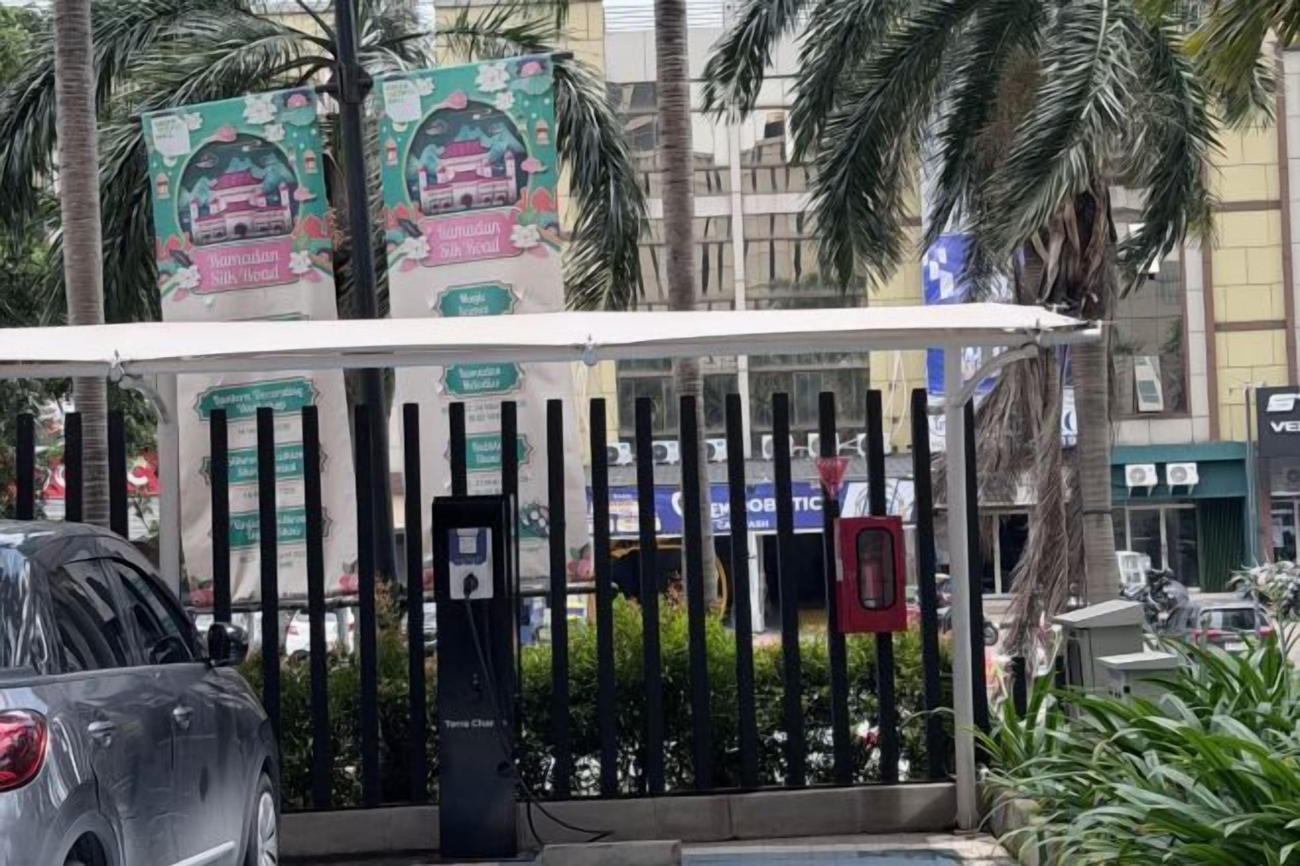 A gated entrance with a covered waiting area, palm trees, and a building with signage in the background.
