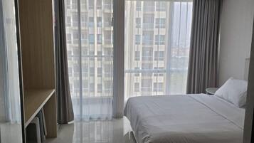 A hotel room with a large bed, a desk, and a view of buildings through sheer curtains.