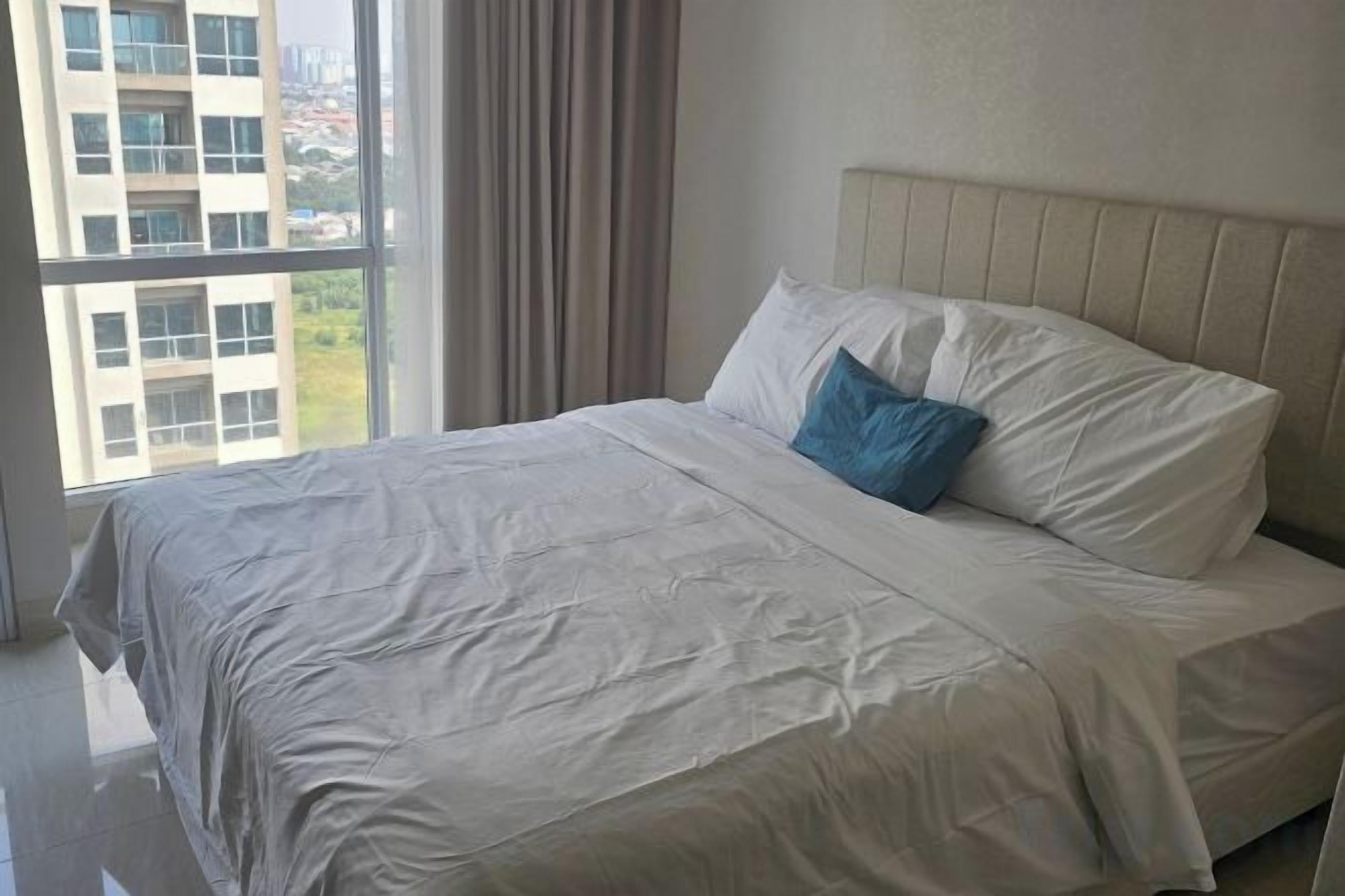 A neatly made bed with a white bedspread and a blue pillow in a room with a large window and a view of buildings outside.