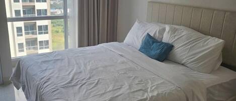 A neatly made bed with a white bedspread and a blue pillow in a room with a large window and a view of buildings outside.