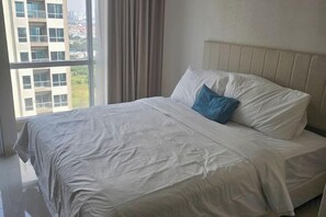 A neatly made bed with a white bedspread and a blue pillow in a room with a large window and a view of buildings outside. - Green Sedayu Apartment Suite by Ellison (West Jakarta)