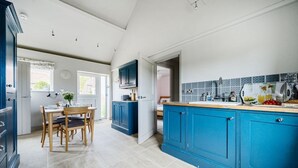 Een moderne keuken met blauwe kastjes, een eethoek met een houten tafel en stoelen, en uitzicht buiten door een raam.