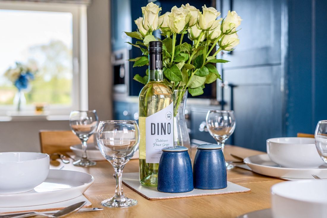 Ett matbord dukat med vita tallrikar, skålar och vinglas, en flaska DINO Trebbiano Pinot Grigio och en vas med vita rosor.
