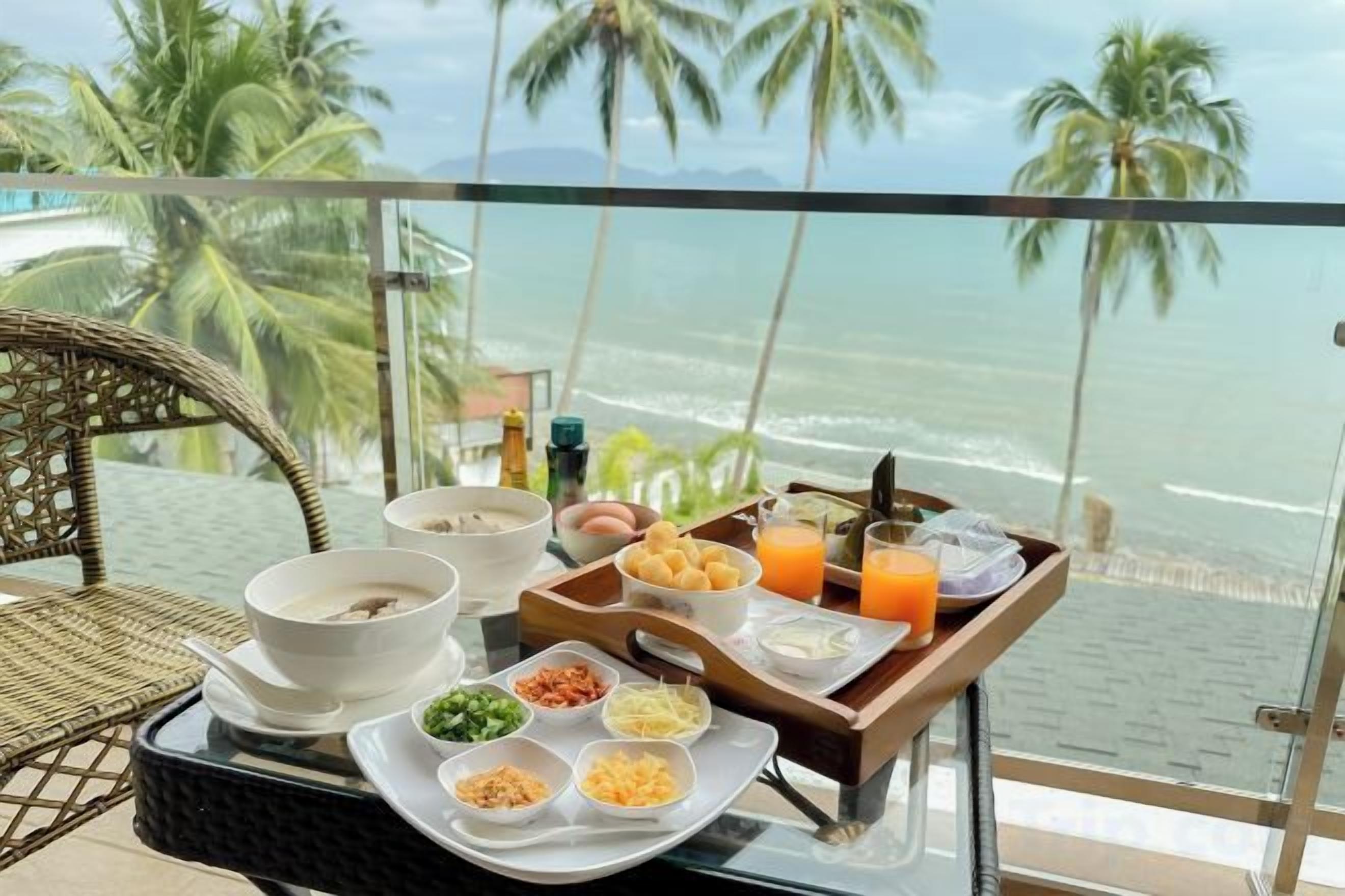 Mesa de pequeno-almoço com bandeja de comida e bebidas numa varanda com vista para o oceano.