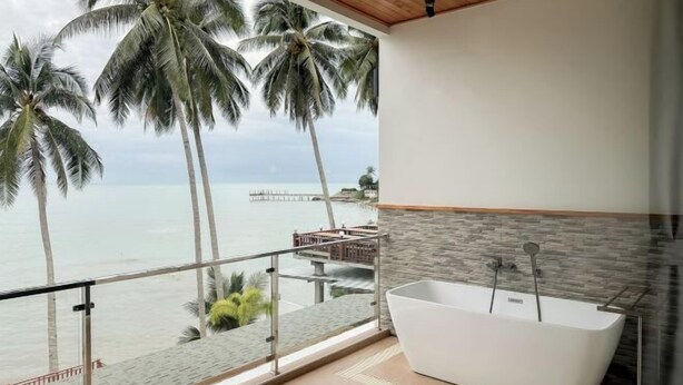 A modern bathroom with a freestanding bathtub, a stone wall, and a view of the ocean and palm trees.