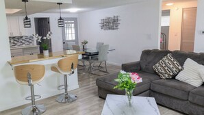 Un soggiorno moderno con un divano grigio, un tavolino da caffè e un'area pranzo con un tavolo in vetro e sedie. È presente una cucina con mobili bianchi e un piano di lavoro in legno.