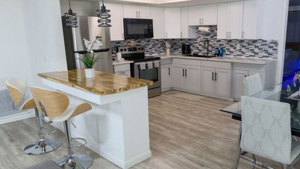 Una cucina moderna con mobili bianchi, un'isola in legno, elettrodomestici in acciaio inossidabile e un tavolo da pranzo in vetro con sgabelli da bar.
