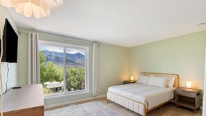 A bedroom with a bed, bedside tables, a desk, a large window with a view, and a ceiling light fixture.