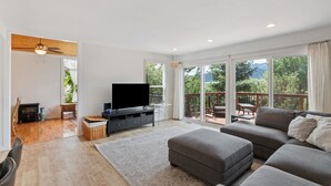 A modern living room with a flat-screen TV, grey sofa, and a balcony with a view of greenery.