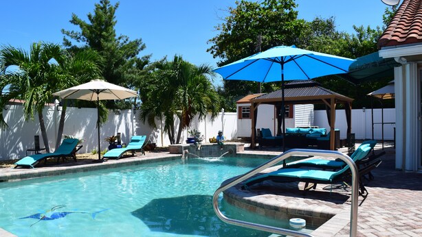 A swimming pool with lounge chairs and umbrellas, surrounded by a white fence and trees.