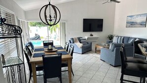 A modern living room with a dining area, a pool view, and a flat-screen TV mounted on the wall.