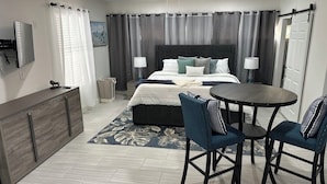 A modern hotel room with a bed, two chairs, a small table, a desk, a TV, and a ceiling fan.