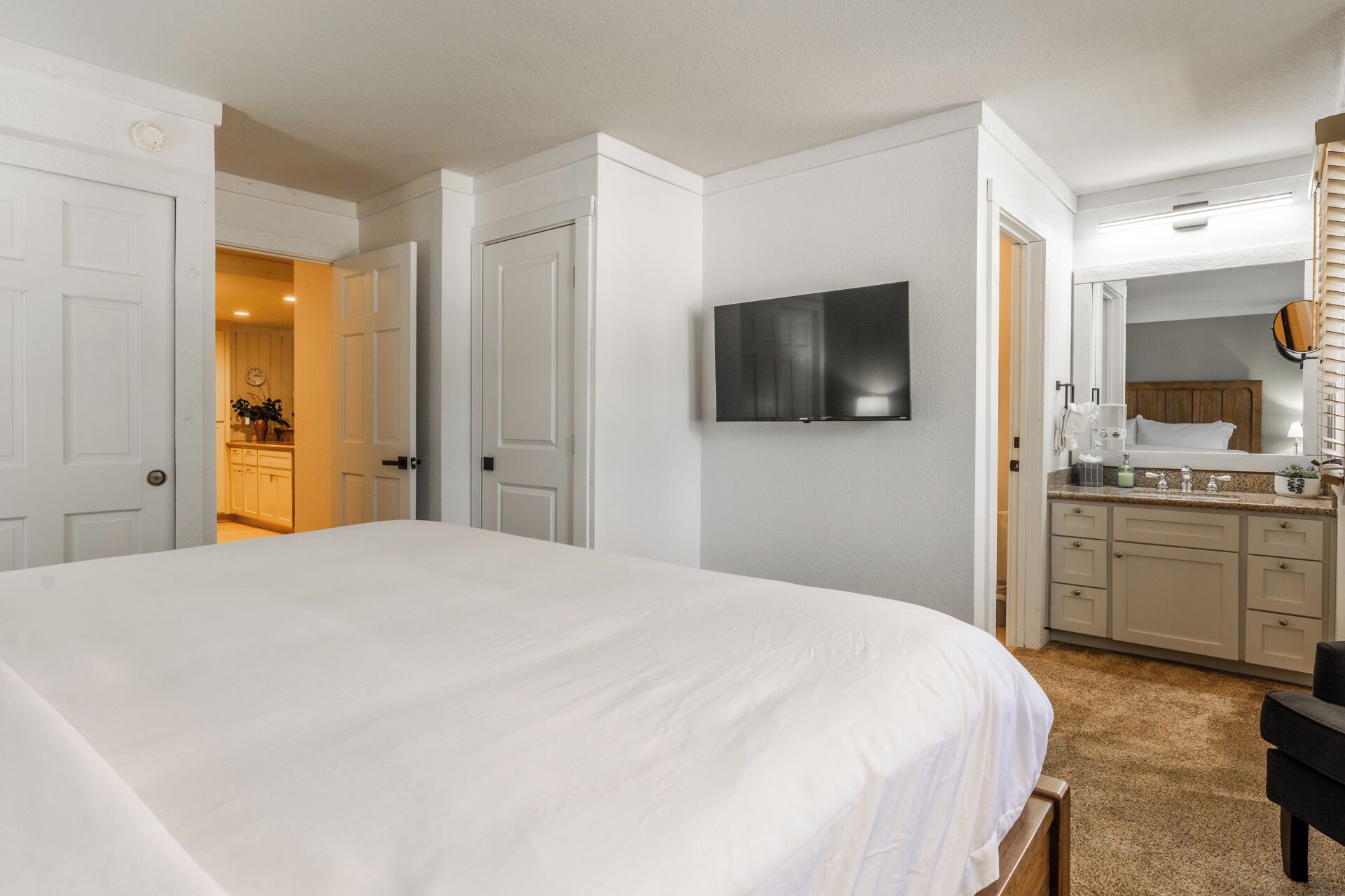 A hotel room with a large bed, a TV mounted on the wall, a bathroom with a mirror, and a kitchenette area.
