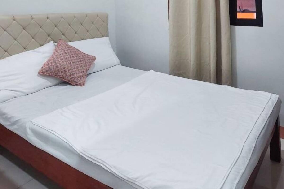 Ein ordentlich bezogenes Bett mit weißer Bettwäsche und einem gemusterten Kissen, ein Knopf-Kopfteil und ein Blick auf einen Vorhang und eine Wand.