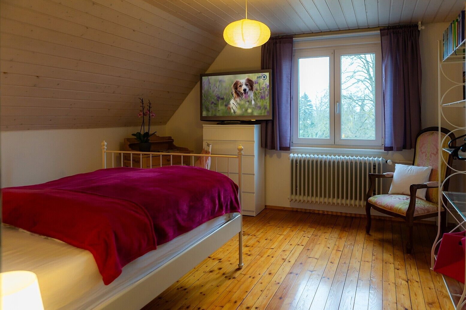 Een slaapkamer met een bed, een radiator, een raam met gordijnen, een stoel en een televisie aan de muur.