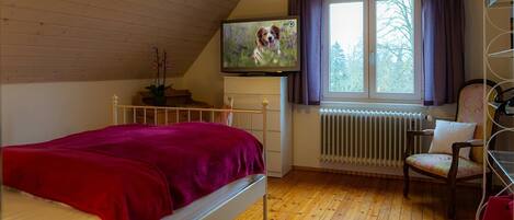 Een slaapkamer met een bed, een radiator, een raam met gordijnen, een stoel en een televisie aan de muur.
