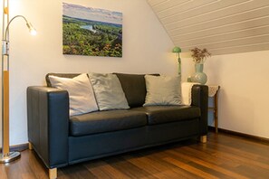 Ein Zimmer mit einem dunkelgrauen Sofa, einer Lampe, einer Vase mit Blumen und einem Landschaftsbild.