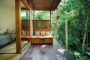 A wooden deck with a sofa and a small table, surrounded by dense greenery. - Ubud Jungle Stay - Mahari by WAHM (Bali)