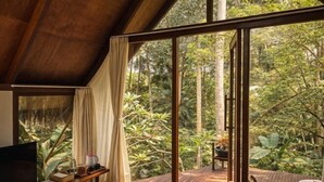 Een kamer met een televisie, een houten tafel en een grote schuifglazen deur die leidt naar een terras met uitzicht op een bos.
