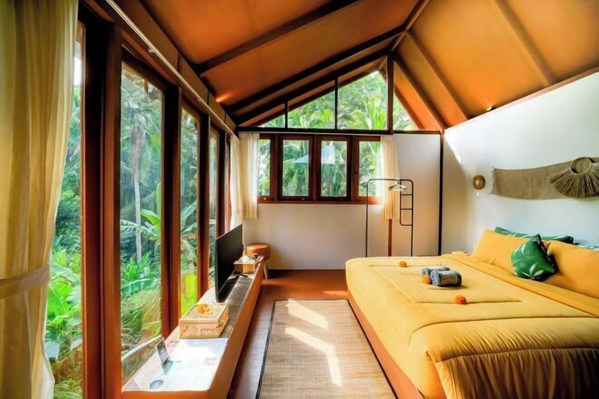 Un dormitorio con cama, escritorio, silla y ventana con vista a la vegetación.