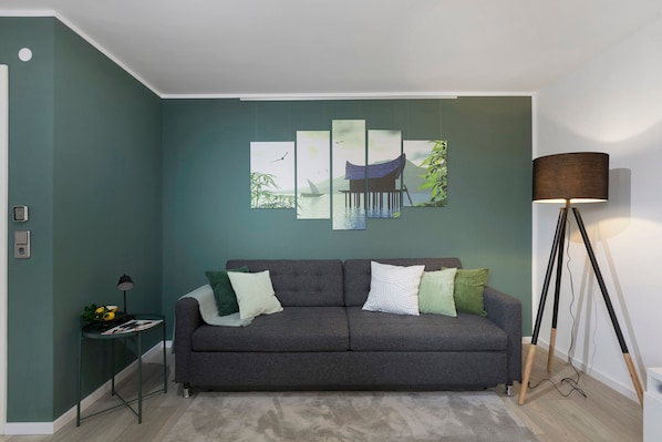 A modern living room with a grey sofa, green cushions, a floor lamp, and a multi-panel artwork on the wall.