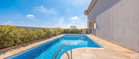 A swimming pool with a metal ladder, surrounded by a stone deck and a low hedge.