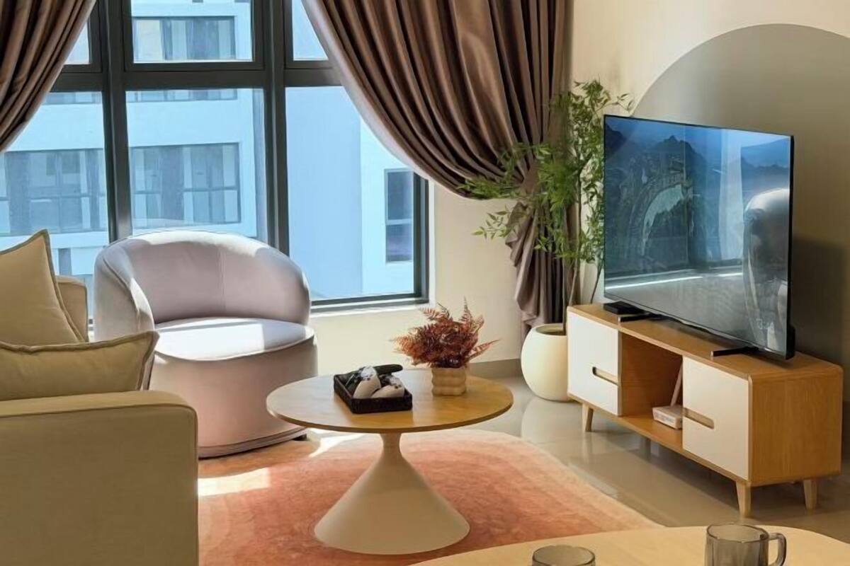 Un salon moderne avec un canapé, une table basse, un téléviseur posé sur un meuble en bois et une grande fenêtre avec des rideaux.