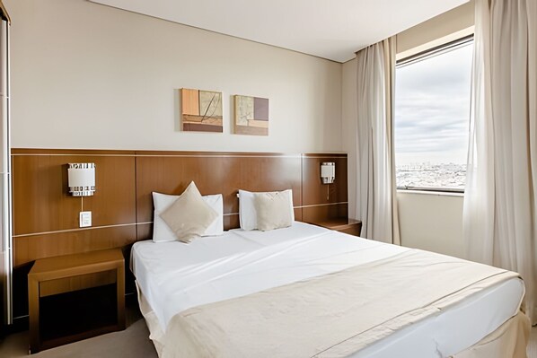 Habitación de hotel con una cama grande, dos lámparas de noche, una mesita de noche y un ventanal con vistas a la ciudad.