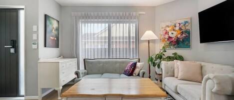 A modern living room with a wooden coffee table, a sofa, a TV mounted on the wall, and a large window with curtains.