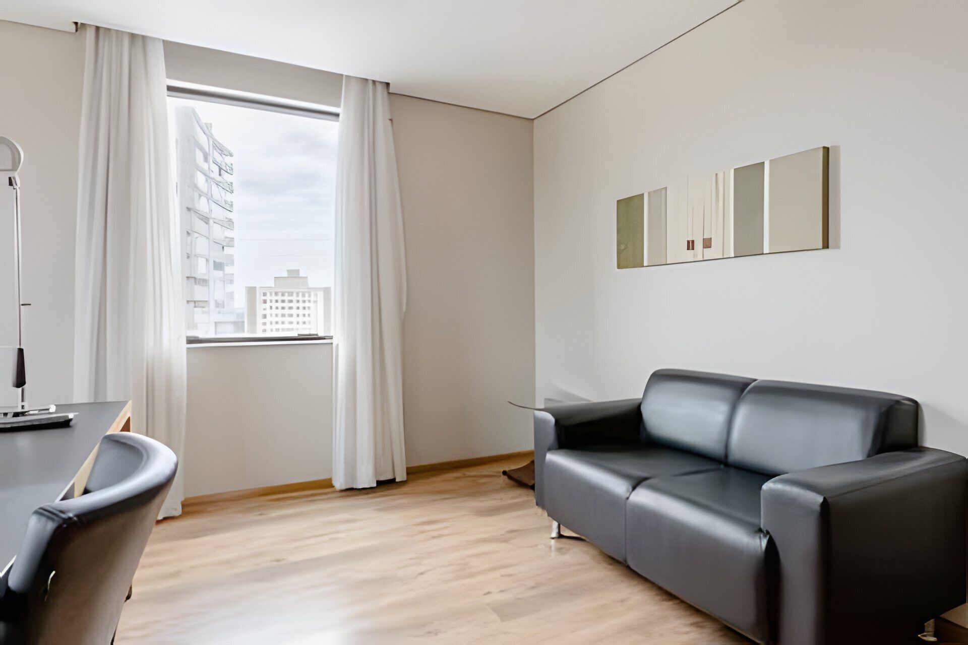 Una sala de estar moderna con un sofá de cuero negro, piso de madera, un ventanal con cortinas blancas y una obra de arte minimalista en la pared.
