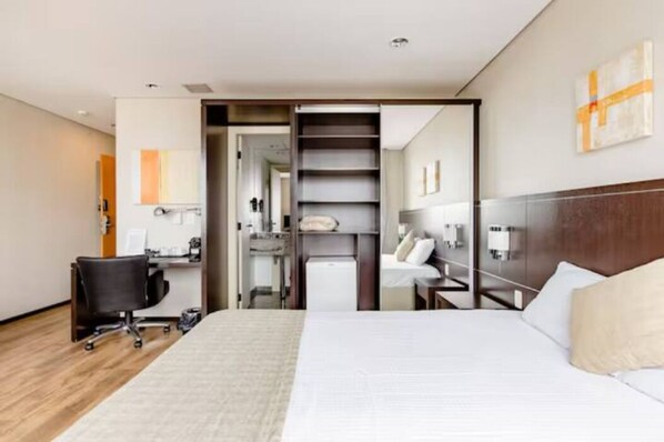 Habitación de hotel con cama, escritorio con silla, armario y una pequeña kitchenette.