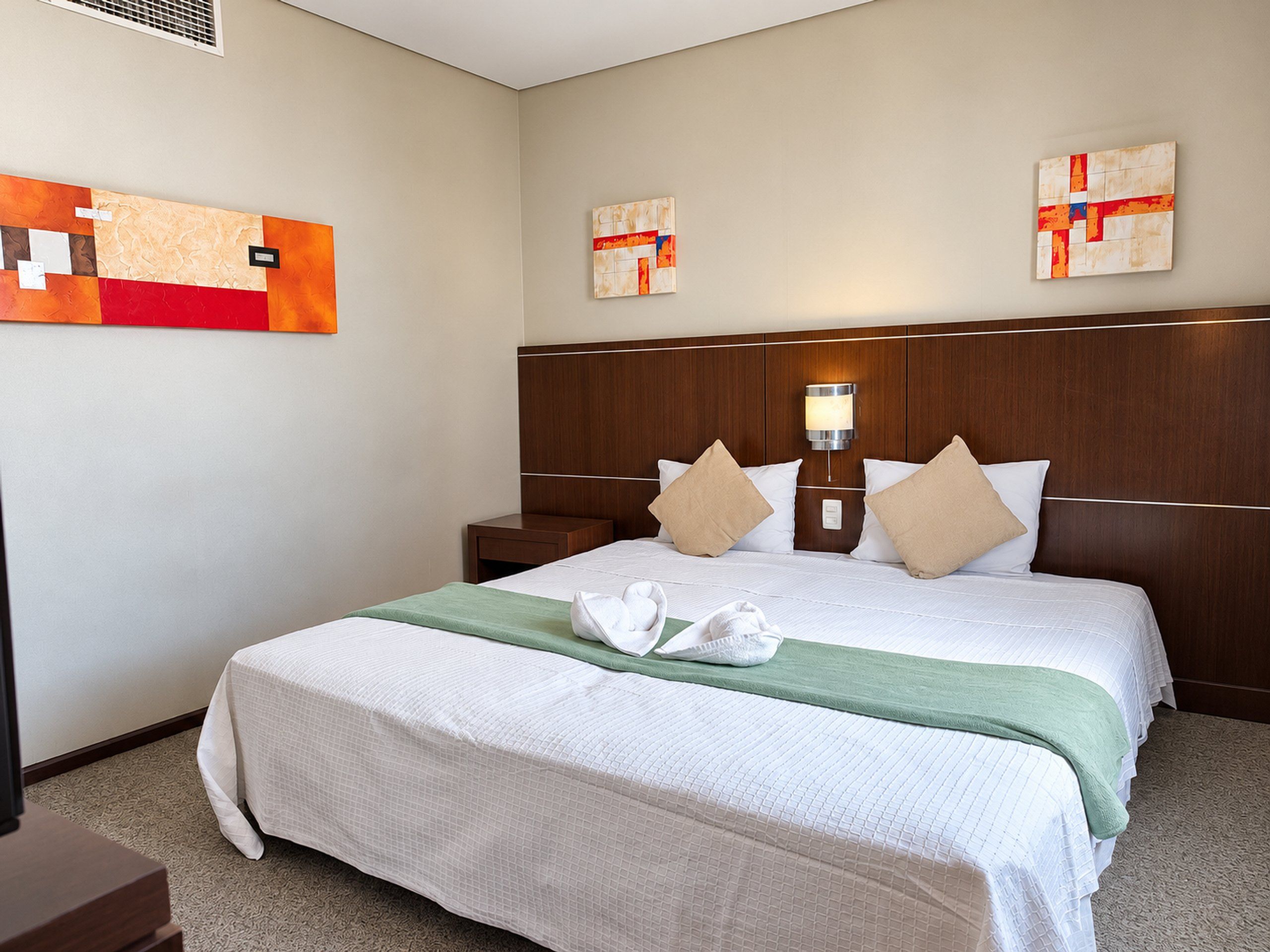 A hotel room with a large bed, two pillows, a nightstand, a wall-mounted lamp, and artwork on the wall.