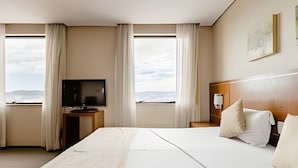 A hotel room with a large bed, a TV on a stand, a nightstand, and a view of the city through the windows. - Apartment with 1 parking space | UN1105 (Osasco)