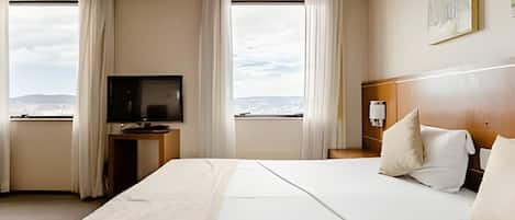 A hotel room with a large bed, a TV on a stand, a nightstand, and a view of the city through the windows.
