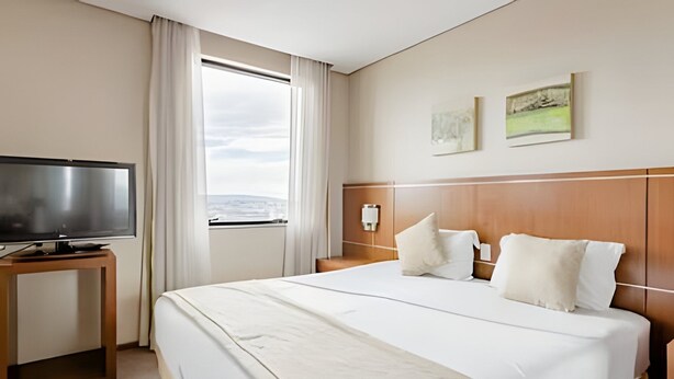 A hotel room with a large bed, a television on a wooden stand, a window with curtains, and two wall-mounted paintings. - Apartment with 1 parking space | UN1105 (Osasco)