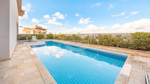A rectangular swimming pool with tiled edges, surrounded by a stone deck, and a view of buildings in the background. - Apartment with 1 parking space | UN1105 (Osasco)
